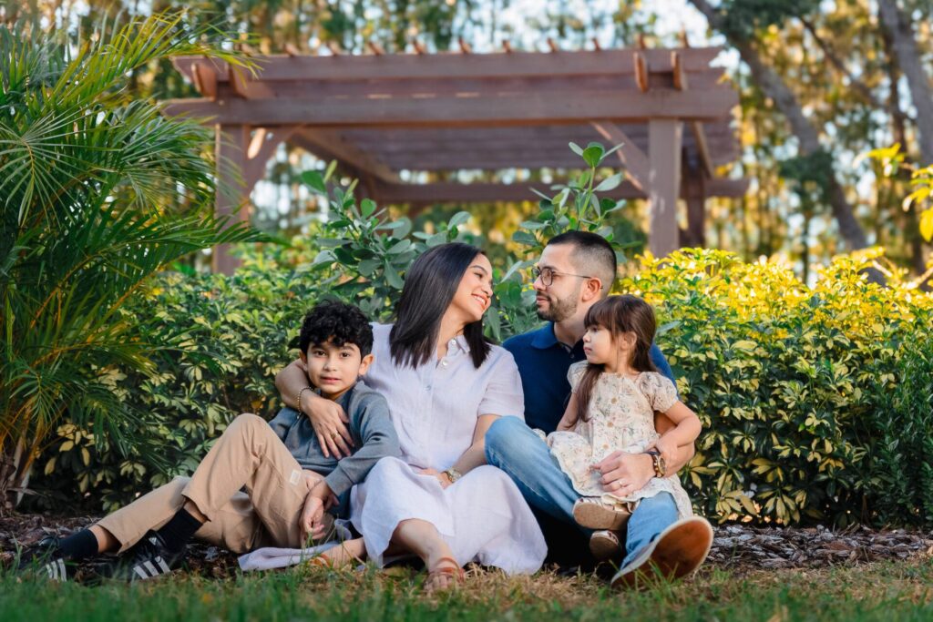 A family of four sitting together in a garden setting, with lush greenery and a wooden pergola in the background. The parents are seated with their young children, creating a warm, joyful atmosphere. Photo taken at Spring Hill by JCenteno Photography. A family of four sitting together in a garden setting, with lush greenery and a wooden pergola in the background. The parents are seated with their young children, creating a warm, joyful atmosphere. Photo taken at Spring Hill by JCenteno Photography.