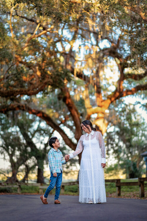 A woman in a white dress holds hands with a young boy in a plaid shirt and jeans, walking on a pathway surrounded by lush trees and natural scenery in Tampa, FL, showcasing professional portrait photography by JCenteno Photography.