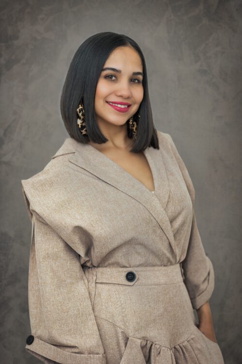 Portrait of a smiling woman with short black hair and leopard print earrings, wearing a beige outfit, in front of a textured gray background. Photography by JCenteno in Tampa, FL.