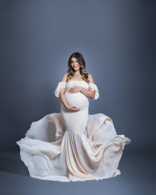 Pregnant woman in an elegant white gown poses for a maternity portrait against a soft gray background, symbolizing professional portrait photography in Tampa, FL.