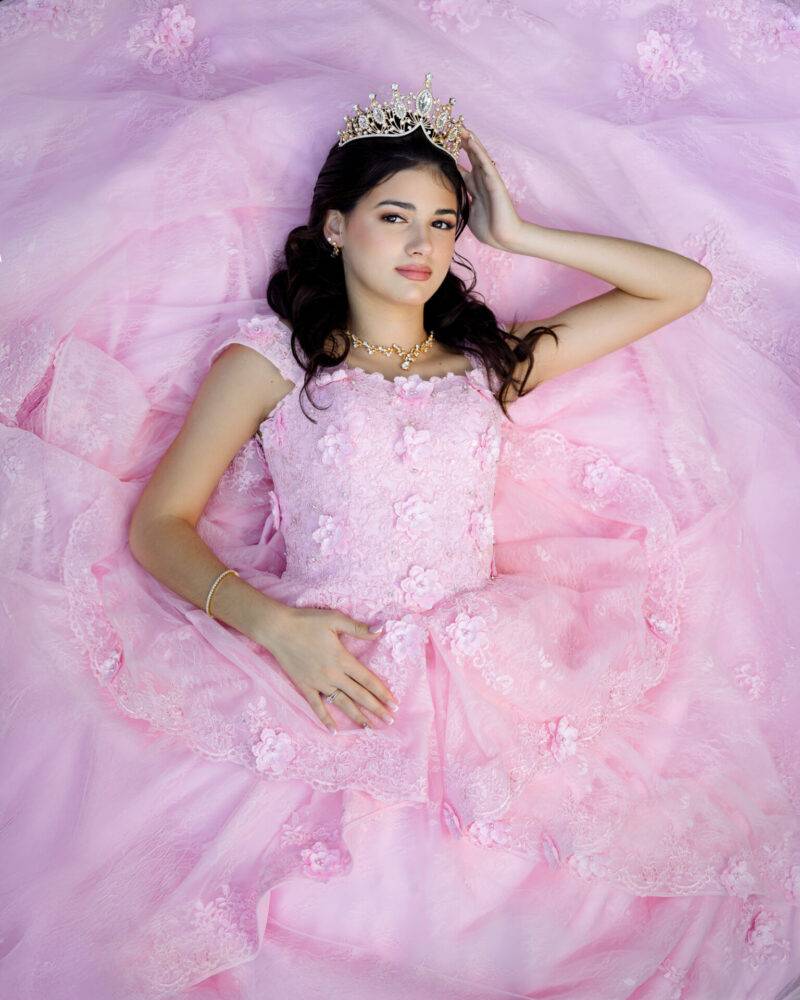 A young woman dressed in an elegant pink quincea&ntilde;era gown lies down, wearing a sparkling crown and delicate jewelry, captured by JCenteno Photography in Tampa, FL.