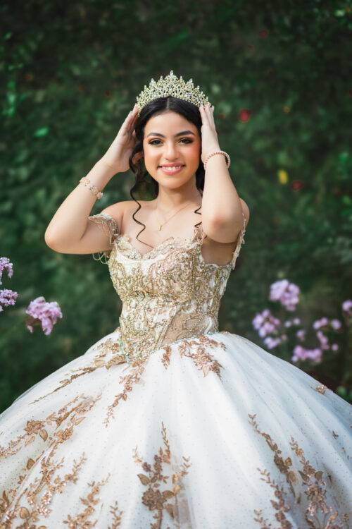 A young woman in an ornate, gold-embroidered quincea&ntilde;era gown poses outdoors. She is adjusting a sparkling tiara on her head, smiling serenely amid a backdrop of blurred green foliage and purple flowers. This portrait showcases expert photography skills, capturing a special occasion in Tampa, FL.