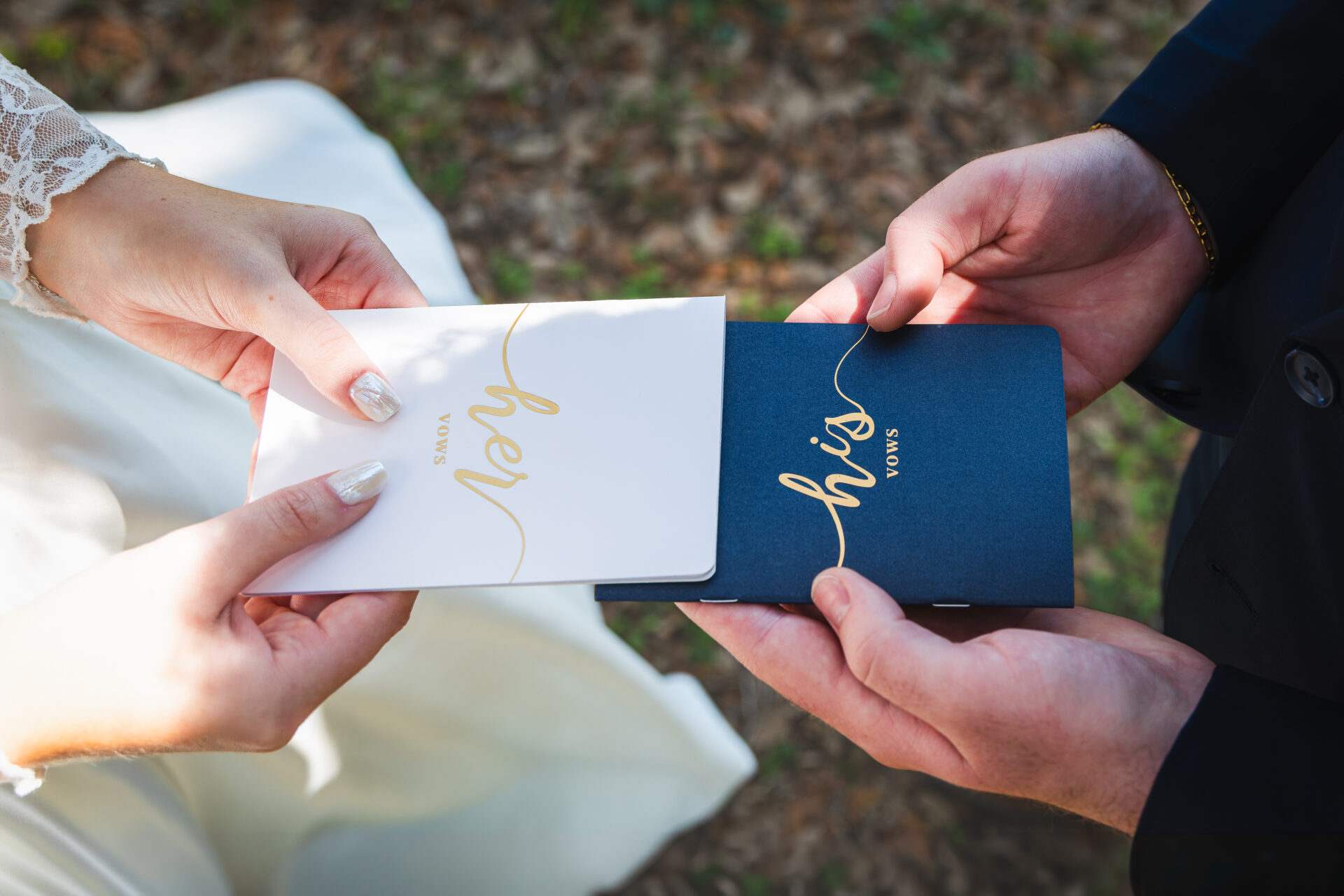 A close-up of a bride and groom holding their vow books during a private exchange. The bride holds a white 'her vows' book with gold lettering, and the groom holds a navy blue 'his vows' book