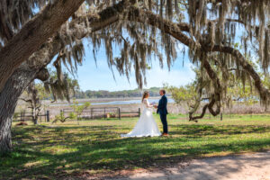 A bride and groom standing face-to-face in a sunny field, exchanging wedding vows during their wedding in Florida. The bride is in a flowing white gown holding a vow book, and the groom is in a navy suit, framed by a massive oak tree with hanging Spanish moss.